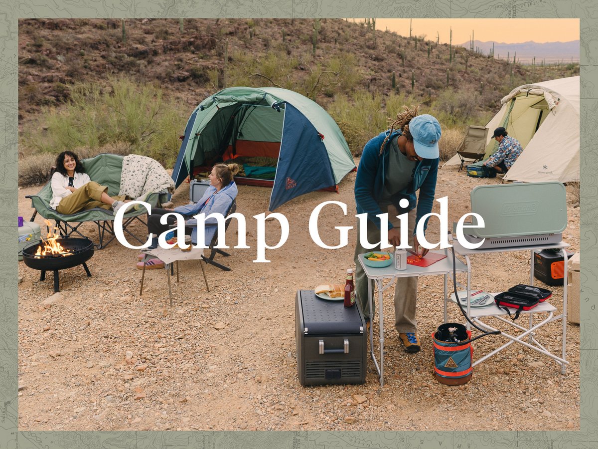 A group hangs out at their desert campsite. Two woman are lounging on comfy camp chairs around a fire. One man is working on dinner at the camp kitchen area. Another man in the back is looking through a bag in his tent.