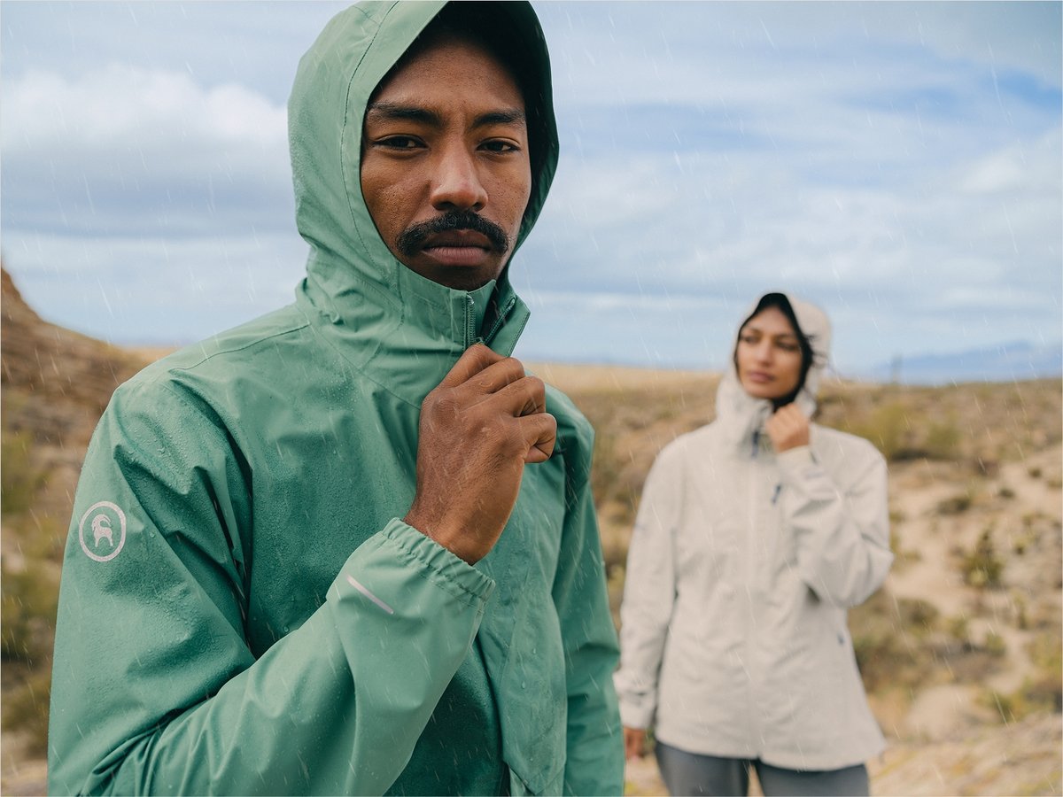 Two people zip up rain jackets as the clouds roll in. Text overlay reads: New Spring Rainwear: Patagonia, The North Face & More, Stay Dry