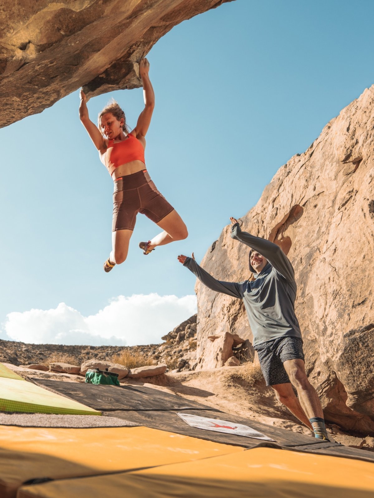 A woman hangs from a cliff midway through a bouldering route. A man stands below her, arms up ready to catch incase she falls. Climbing pads are spread underneath her.