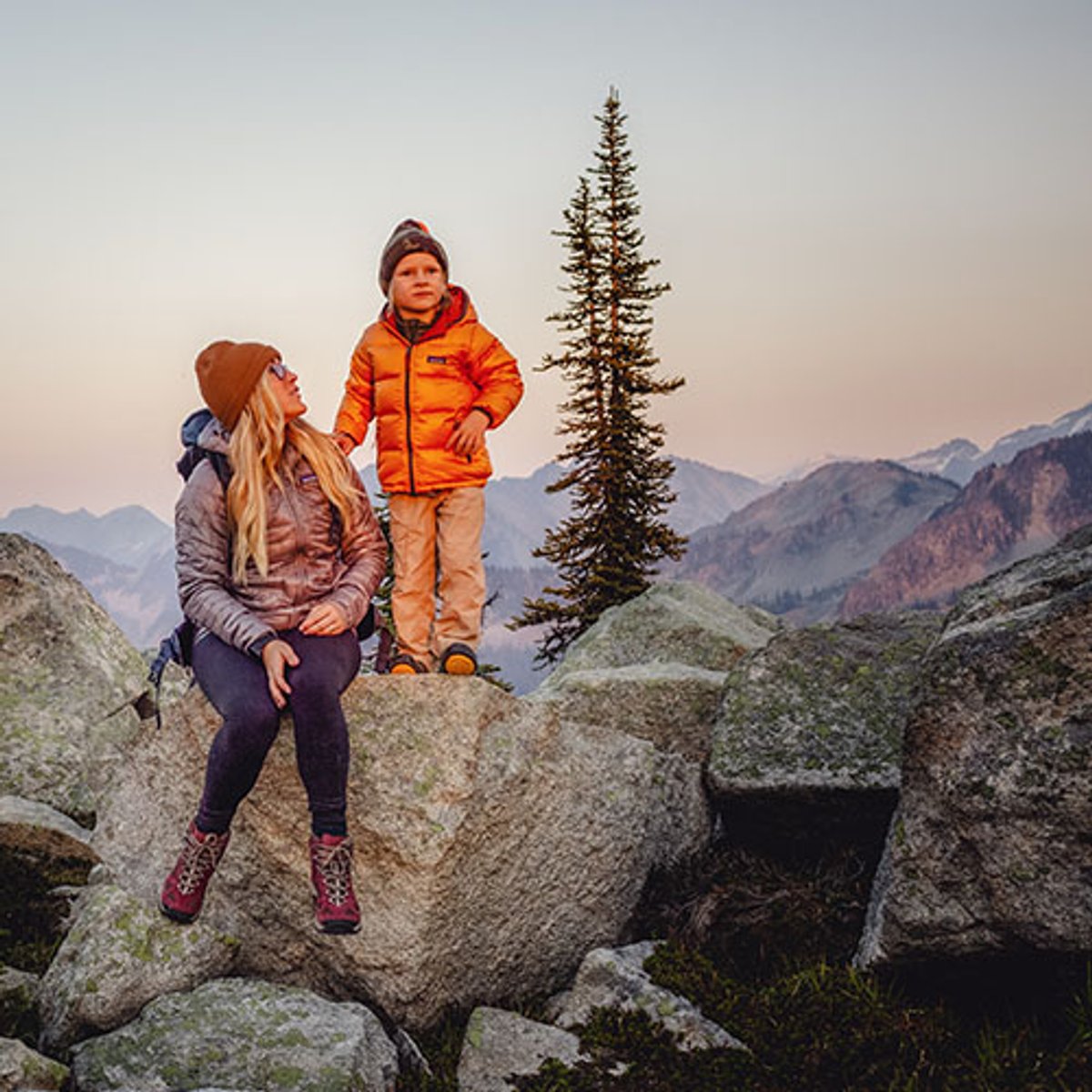 a parent and child sit on a rock at the top of a mountain, wearing coats and hats