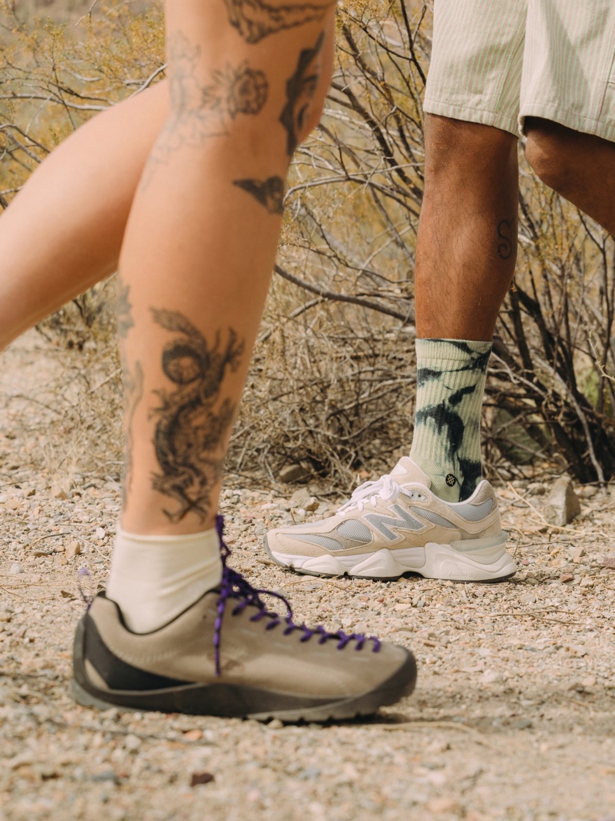 Two legs from different people walking past each other feature this season’s top footwear. One leg is covered in tattoos. They are walking on a desert landscape.
