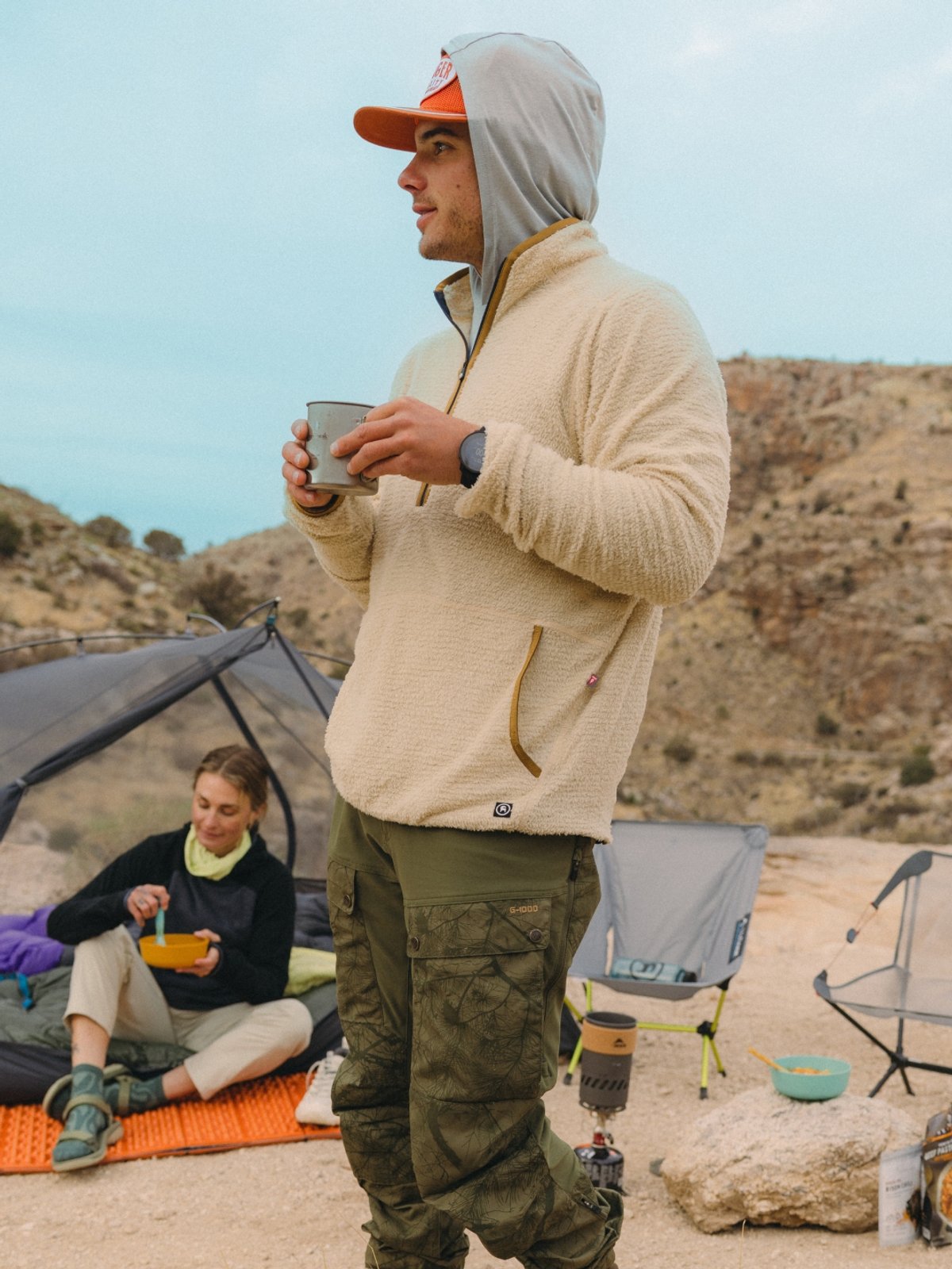 A man in a fleece sweater and green pants holds a cup of coffee. In the background, a woman is in a tent eating out of a bowl.  They are desert camping, sandy hill with shrubs rise behind them.