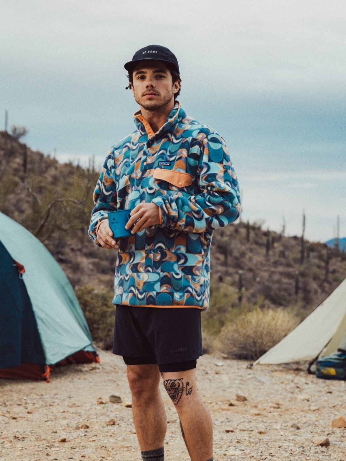 A man in a jazzy Patagonia sweater stands in a desert landscape between two tents. Saguaros are silhouetted on the dark horizon.