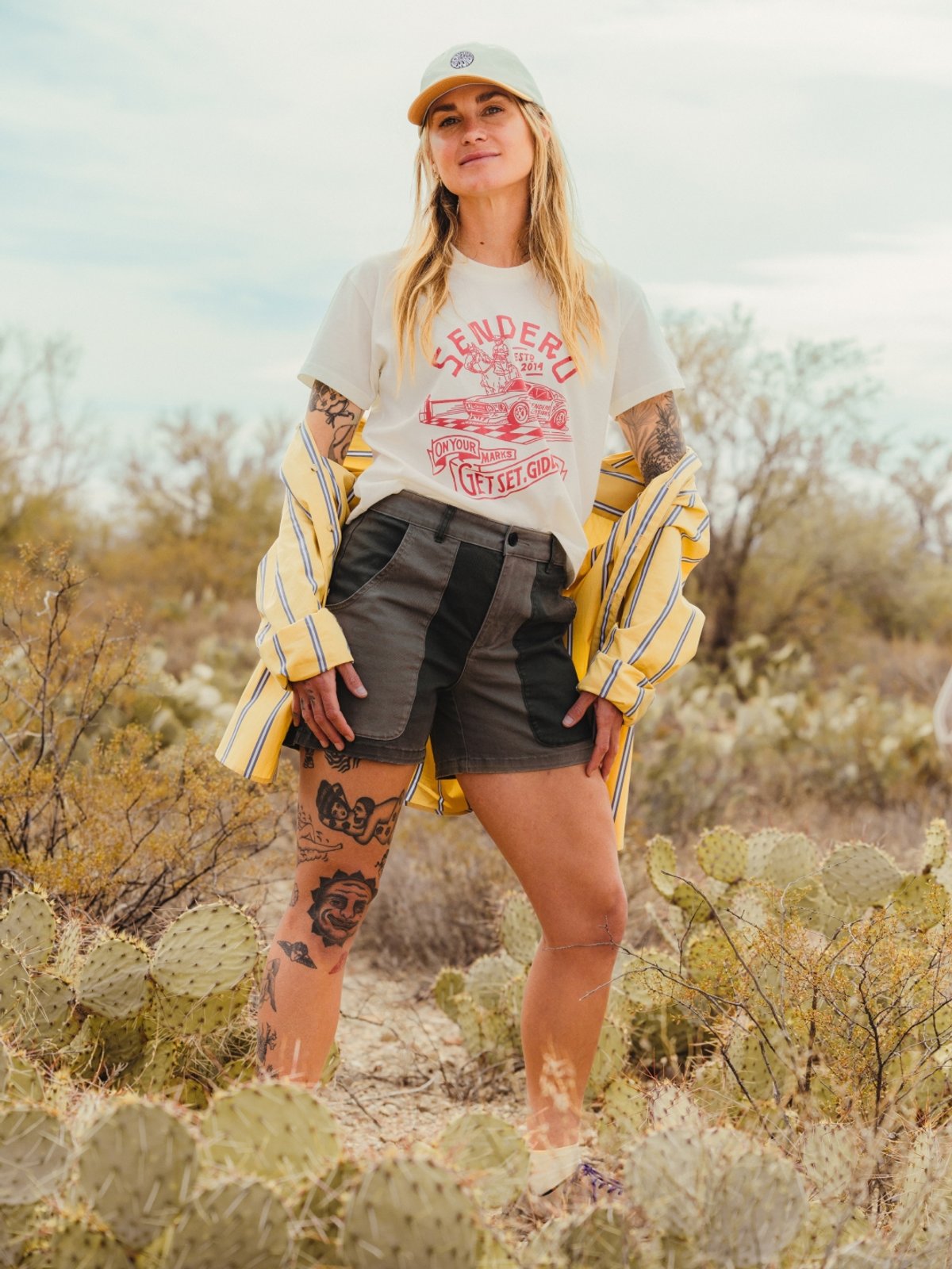 A woman in a t-shirt and shorts stands in a desert landscape among prickly pear cacti.